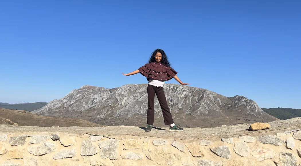 A girl standing with her arms open on a stone wall in front of a rocky mountain