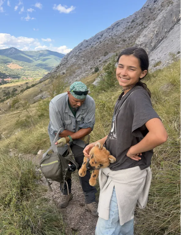 An adult with a backpack and a smiling girl holding a toy fox on a mountain trail