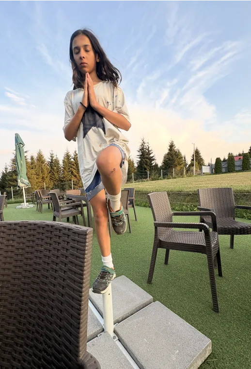 A girl balancing on one leg with hands together on an outdoor terrace, trees and evening sky behind her