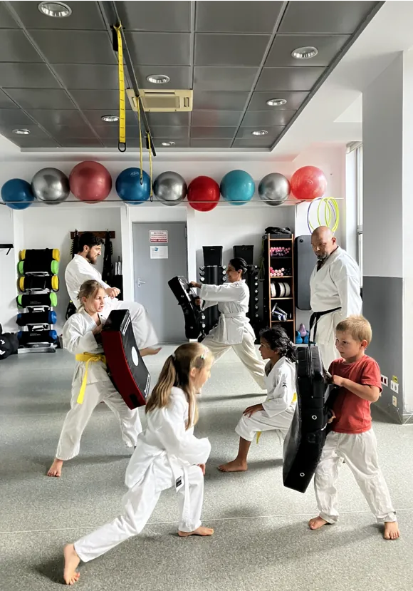Parents and children in white karate uniforms practising kicks on pads in a training hall